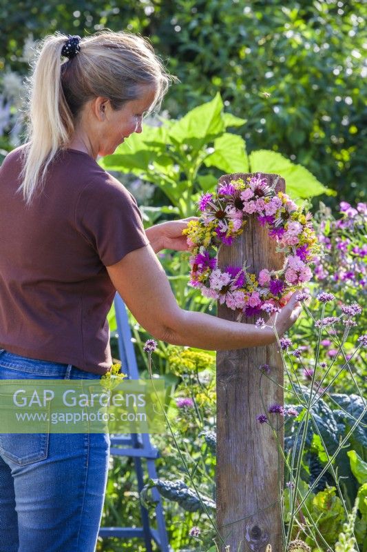 A woman hangs a mostly pink wreath made of  Echinacea, Rosa, Fennel, Monarda and Veronicastrum on a wooden stand