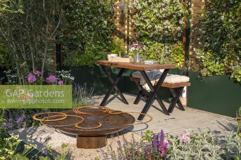 An urban courtyard garden with wooden bench and table, corten steel water feature, raised bed containers planted with Trachelospermum jasminoides climbing over hazel fencing screens