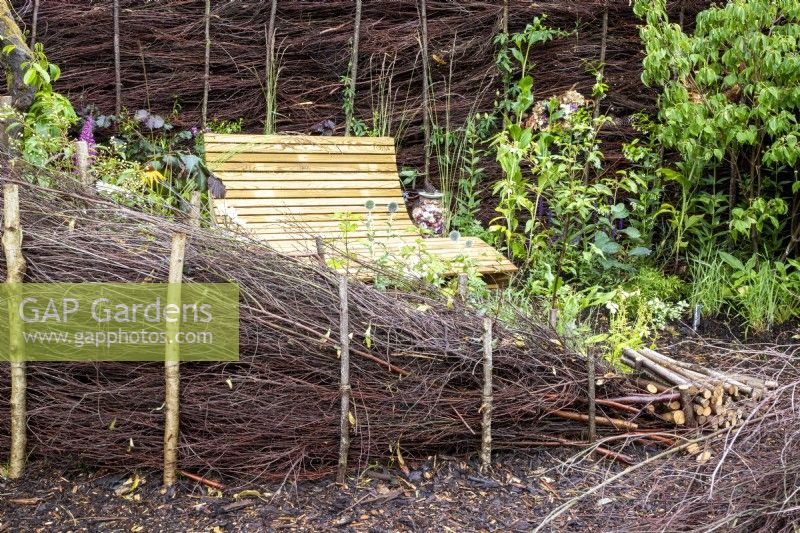 A dead hedge made from tree prunings with a wooden bench in an enclosed area 
