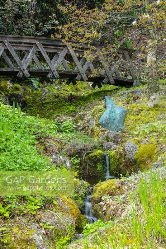 An arched wooden bridge over a waterfall and bronze sculpture 'Rapture'.