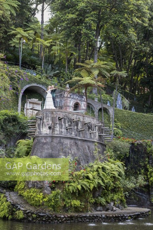 A newly married couple stand, Titanic style, on top of the Fortress, a water feature, set within the Central Lake. Monte Palace Gardens, Madeira. August. Summer