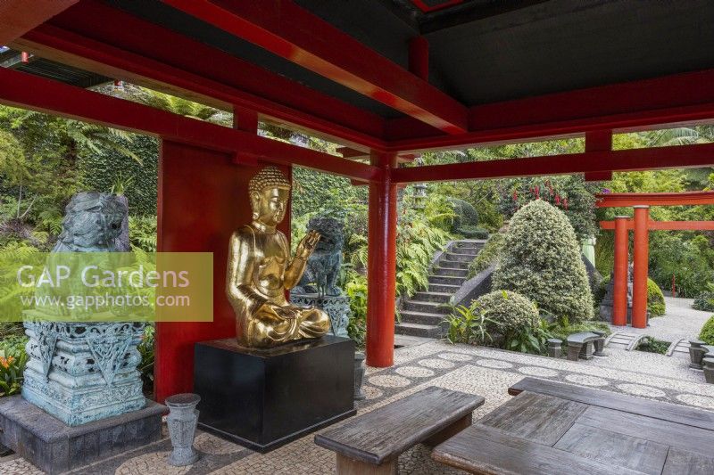 A large seated gold Buddha within a red Oriental style pagoda with Foo dog statues, wooden furniture and a tropical garden in the background. Monte Palace Garden, Madeira. August. 