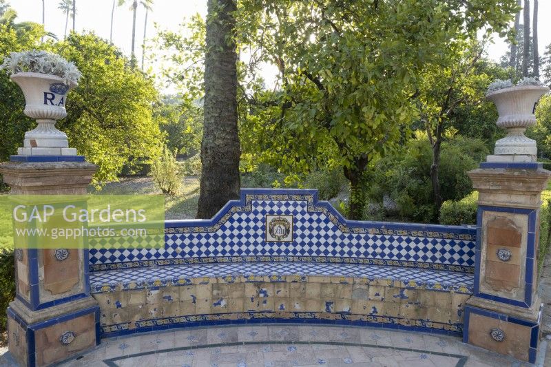 A chequered tiled and curved bench with pillars at the ends and ornate urns set on the pillars. Real Alcazar Palace gardens, Seville. Spain. September. 