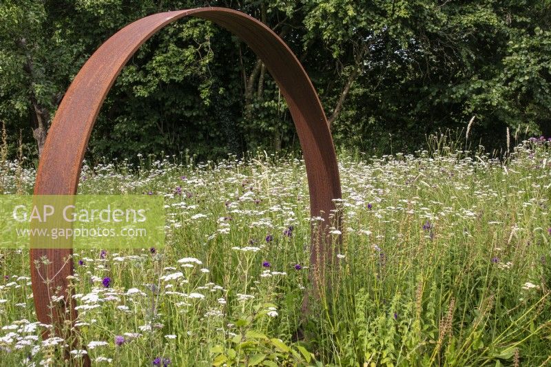 A Corten steel moon gate stands in a perennial meadow with Achillea millefolium.