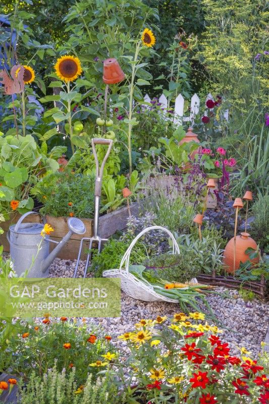 A basket of picked vegetables in front of a bed of mixed herbs and raised beds full of growing vegetables and annual flowers.