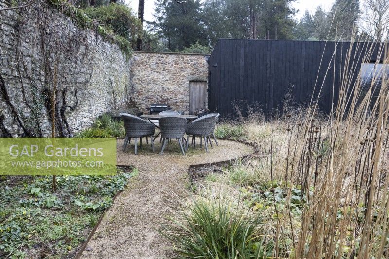 A chipped path leads to a circular section with a round rattan table and chairs on. Various grasses and plants are to the right. Winter. Italian Garden at Great Ambrook. 