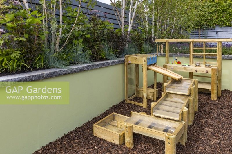 A small play corner with a wooden mud pie kitchen and water run on floor of pine bark. The raised border with Betula utilis 'Multi Stem', Sambucus nigra 'Black lace' and Festuca glauca 'Elijah Blue'. June. Designer: Colm Carty, Bord Bia Bloom 2023 