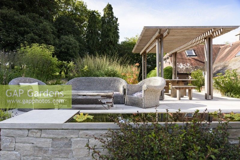 Terrace in a country garden in July with wicker furniture and a wooden gazebo seen beyond a water feature