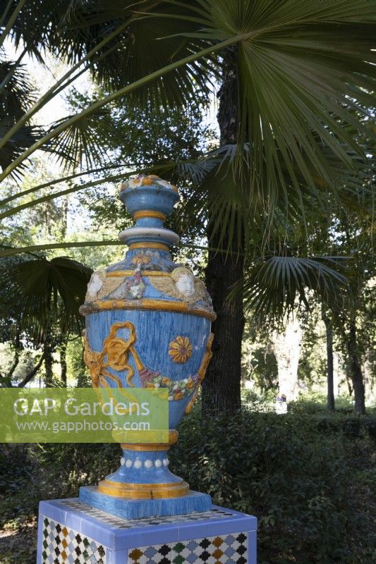 A colourful, glazed urn, on top of a plinth. Various shrubs and trees are in the background.  Parque de Maria Luisa, Seville, Spain. September