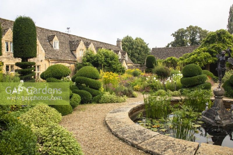 View across The Flower Garden at The Manor, Little Compton, with perennials, topiary and a circular pond with fountain.