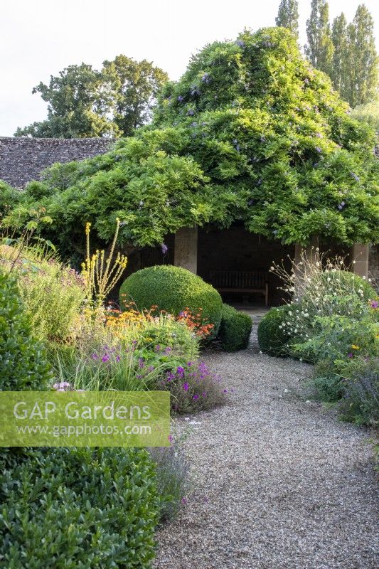 A gravel path leads past topiary and flowerbeds to a wisteria-covered summer house in the Flower Garden at The Manor, Little Compton.