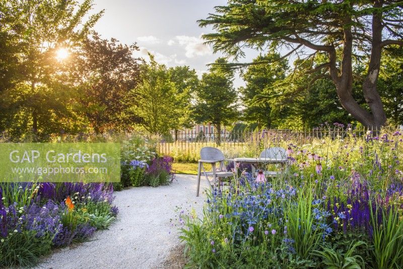 Gravel path leading to seating area with wooden table and two chairs. Planting includes RHS Iconic Horticultural Hero Garden, Designer: Carol Klein, RHS Hampton Court Palace Garden Festival 2023