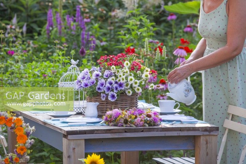 A woman pours tea into a cup on a table with a central floral arrangement that includes Surfinia, Verbena and Scaeola in a wicker container.
