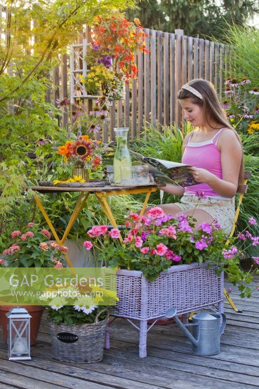 Container plantings on the terrace with Impatiens and Pelargonium, a flower arrangement with a cold drink on the table and a girl enjoys reading a gardening magazine.