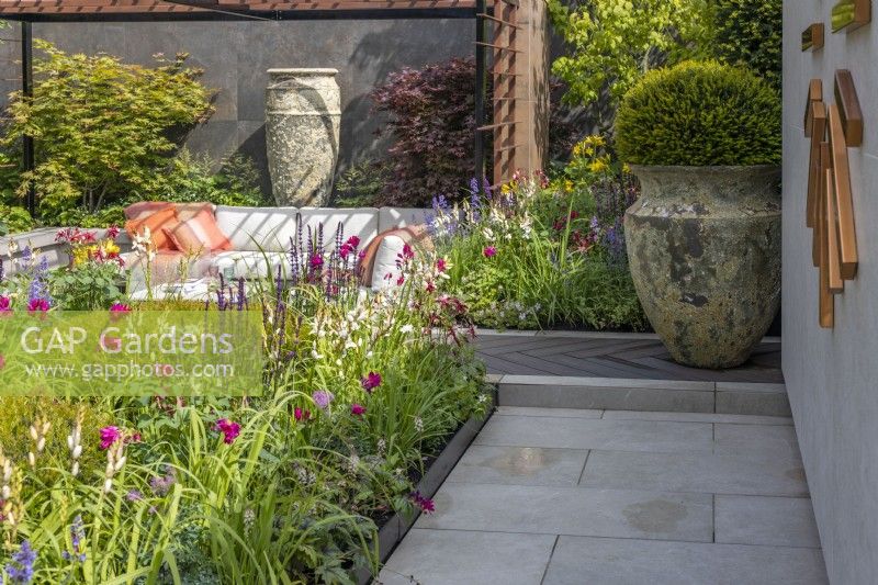 A colourful perennial bed along modern paving slabs and next to a large ceramic vase planted with a yew ball in a contemporary garden. Garden: Looms  and  Blooms, Designer: Kevin Dennis, Bord Bia Bloom 2023