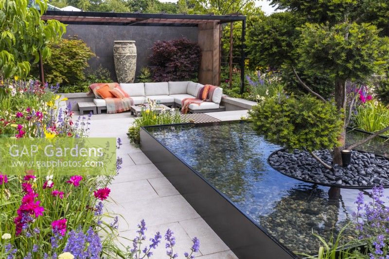 A view of the modern garden with a seating area under a steel pergola, colourful flower beds and a water feature with a trimmed yew tree in the middle. Designer: Kevin Dennis, Bord Bia Bloom 2023 