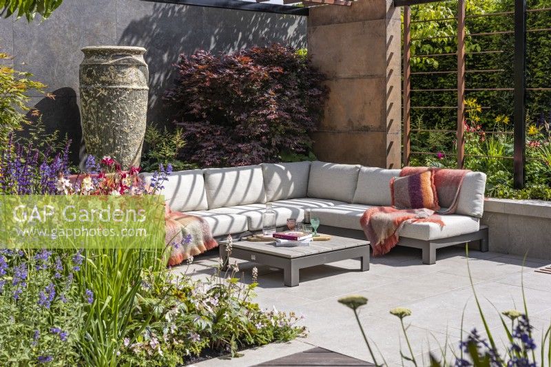 A sunken seating area with a modern corner sofa, low table, large ceramic vase and maple tree surrounded by colourful perennial beds. A steel pergola creates a mottled shade. Designer: Kevin Dennis, Bord Bia Bloom 2023 