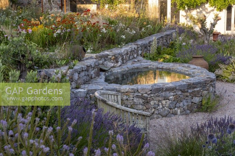 Small circular pond with stone sides in drought tolerant garden planted with Lavender, Verbena hastata and Heleniums