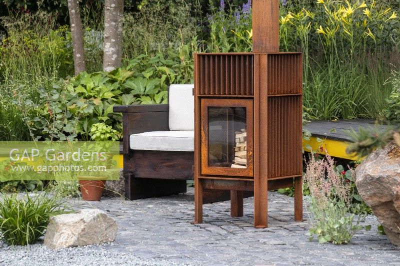 A seating area with chair and metal outdoor wood burning stove set on reclaimed granite setts Hurtigruten: The Relation-Ship Garden, Max Parker-Smith, RHS Hampton Court Palace Garden Festival