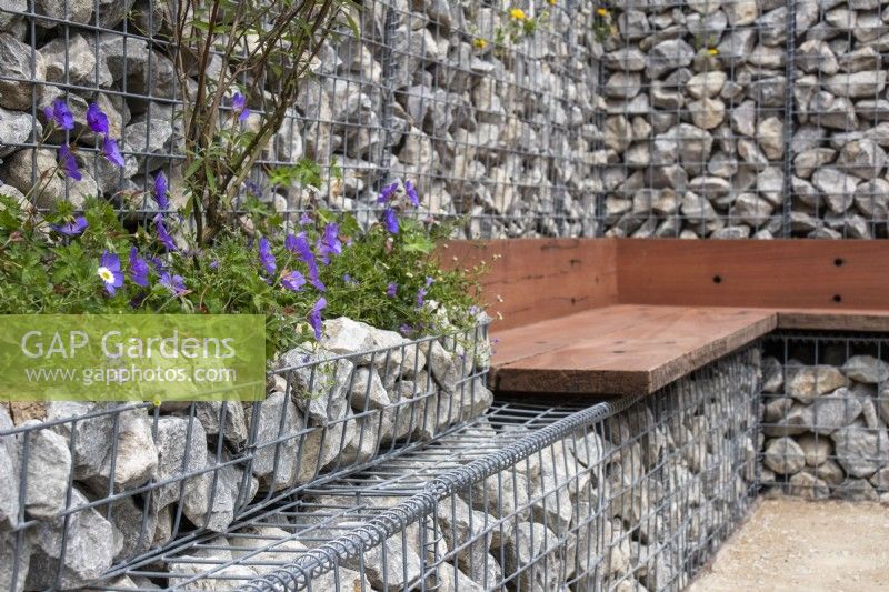 A seating area using reclaimed wood against a boundary wall made of gabions filled with stone offcuts. Geranium 'Johnston Blue' is planted into the gabions - - Caroline and Peter Clayton - Get Started Gardens - Nurturing Nature in the City, RHS Hampton Court Palace Garden Festival.