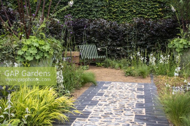 Grey brick and cobbled path leading to a seating area with a metal chair. Planting in borders and a large container includes Halonechloa macra and Stipa tenuissima  - designer Lucy Taylor - The Traditional Townhouse Garden -  RHS Hampton Court Palace Garden Festival.