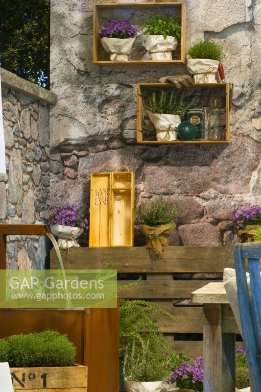 Rusted corten steel water tank by the wall stone, and herbs in containers and on pallet,  crates with herbs hanging on the wall in contemporary Italian courtyard.