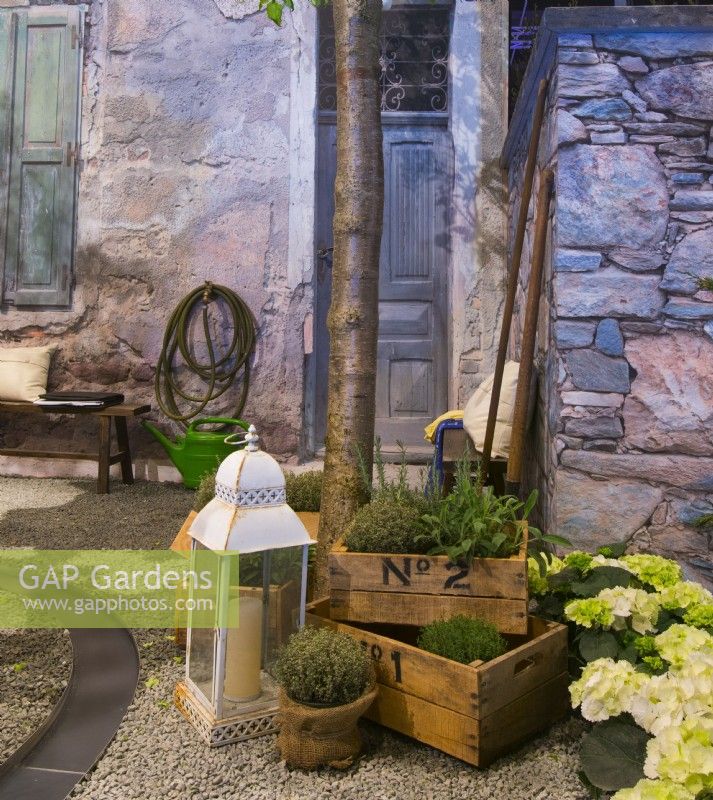 Display of herbs and Hydrangea in containers and lantern in front of the stone house of contemporary Italian courtyard. A water canal running on the gravel surface. 