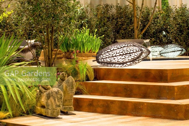 View of steps made from natural stone treads with corten steel risers. African sculptures in foreground and a decorative floor lamp. Designer: Vetschpartner, Berger Gartenbau and Livingdreams. Giardina-Zurich, Swiss.
