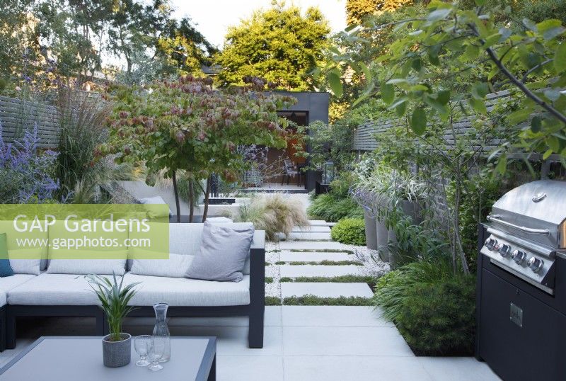 View of the seating area and outdoor kitchen with multi stem Parrotia persica planted behind the sofa, by the barbecue, Pinus mugo and Amelanchier canadensis with Thymus praecox 'Albiflorus' running between the paving stones leading to the zinc clad building.