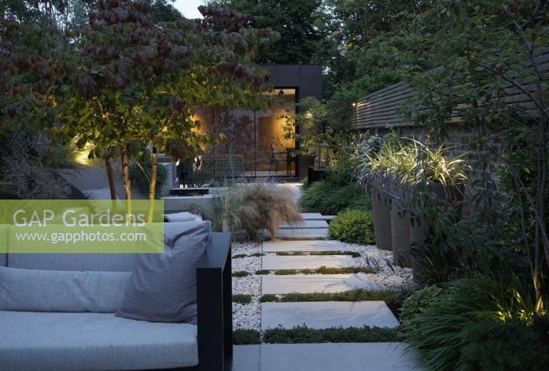 View of this city garden in the evening with outdoor lighting dotted around the planting and the path of sanstone slabs leading to the zinc clad building 