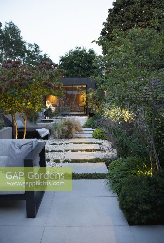 View of this city garden in the evening with outdoor lighting dotted around the planting and the path of sandstone slabs leading to the zinc clad building 