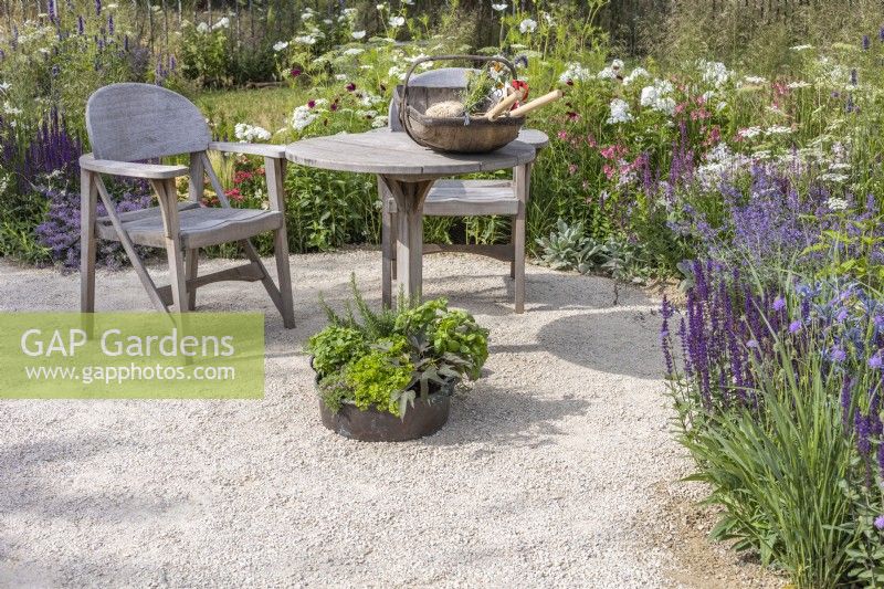 A seating area on a gravel surface with wooden table, two chairs and inset herb planter surrounded by summer border planted with perennials and ornamental grasses. RHS Iconic Horticultural Hero Garden, Designer: Carol Klein, RHS Hampton Court Palace Garden Festival 2023