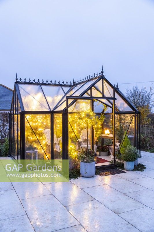 Large greenhouse furnished and decorated with recycled plastic chairs, fairy lights and mixed planting on patio