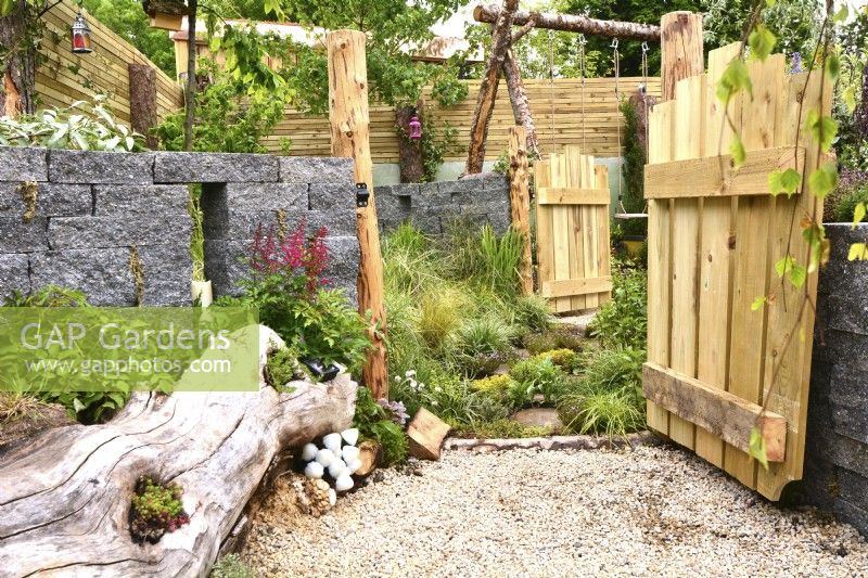 Open wooden a gate leads into a grass area for dogs, alongside a large tree stump lying on a gravel surface used as a flowerbed and seating area next to a decorative Connemara wall system  in a woodland inspired garden. Planted with Astilbe, fungi, grasses. June
Designer: Mary Anne Farenden. Bord Bia Bloom, Super Garden, Dublin, Ireland.


