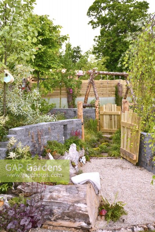 Rustic woodland inspired garden with a large tree stump used as a flowerbed and seating area, open wooden gates leading to different area. Planted with  Astilbe koreana,  Pyrus salicifolia, Betula pendula. June
Designer: Mary Anne Farenden. Bord Bia Bloom, Super Garden, Dublin, Ireland.