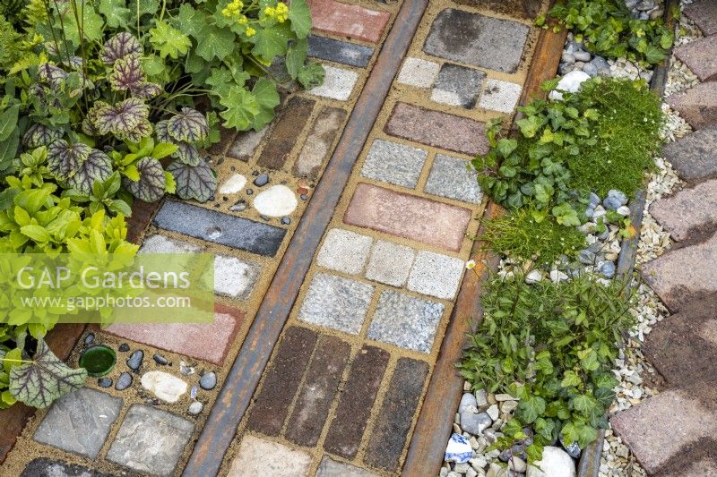 Varied materials: reclaimed cobbles, tiles, bricks, rusted rails and stones used as garden mosaic paving between strip beds planted with Heuchera, Hedera helix and Alchemilla mollis. Designer: Nicola Haines, Citroen Power of One at Bord Bia Bloom Dublin 2023.