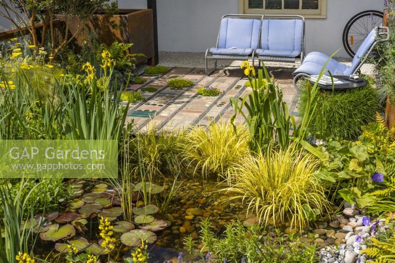 Urban front garden with pond and seating area. Recycled seats are from an old Citroen car model. Mosaic terrace surface made of various reclaimed natural materials. Pond with Nymphaea surrounded by Acorus gramineus 'Tail' and Iris pseudacorus. 
