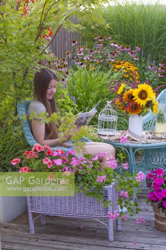 A young woman enjoys reading a magazine on a decked summer terrace with containers planted with pelargoniums.