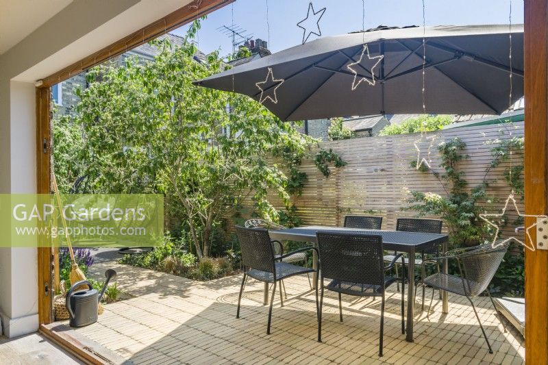 A small sun filled patio garden designed as an outdoor living space with large parasol, garden table and chairs and a pair of hanging seats. June.