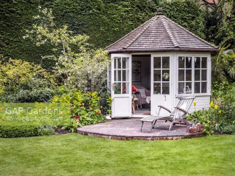 A small wooden summerhouse and patio with mixed borders including Tulipa 'Brown Sugar', 'General de Wet', 'Sunny Prince', Smyrnium perfoliatum and Rubus deliciosus
