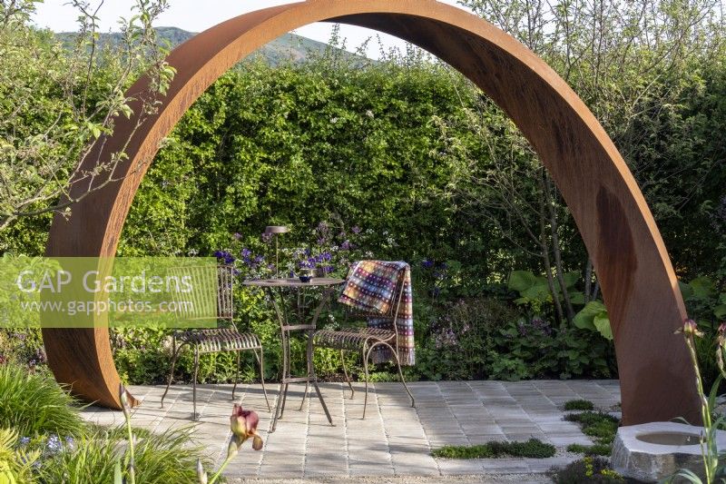 Metal table and chairs on paving under a Corten steel arch  - 'It Doesn't Have to Cost the Earth' - designer Michael Lote - RHS Malvern Spring Festival 2024