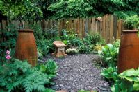 Small patio garden with slate chippings, foliage planting, rustic wooden fence, and terracotta urns and seat