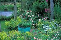 View of wrought iron bench amongst Alchemilla mollis - Lady's Mantle, pink roses and peonies with a lake in the background.