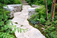 A path leading to curved stone seats surrounded by shady woodland planting including Betula papyrifera, Phlox divaricata, Aquilegia canadensis and ferns 