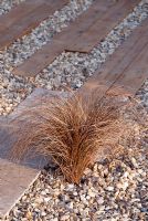 Carex flagellifera planted in gravel with wooden and stone paving in 'The Homebase Living Room' garden 