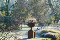 Terracotta urn on a plinth on a frosty winter's morning in John Massey's garden. Carex comans 'Bronze form'. Grasses border, lawn and trees beyond. 