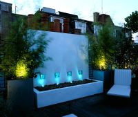 Roof garden lit up at night with decking, white water feature, chairs and galvanised pots planted with bamboos
