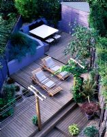 View of split level garden with pergola with vine, sun loungers, decking, shed, raised bed with blue grasses, shell mulch, Hosta and Acer in pots