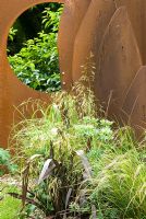 Phormium, Eryngium giganteum 'Miss Willmott's Ghost' and grasses set against a sculpture made from a rusting plough and metal panel by Lucy Redman. Prunus lusitanica - Portugal laurel seen through circular hole in rusting panel