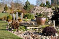 A sunny spring day in John Massey's garden with Prunus incisa 'Kojo-no-mai' flowering in a large pot on the deck by the pond at Ashwood Nurseries. Decorative duck ornaments and slate pillars